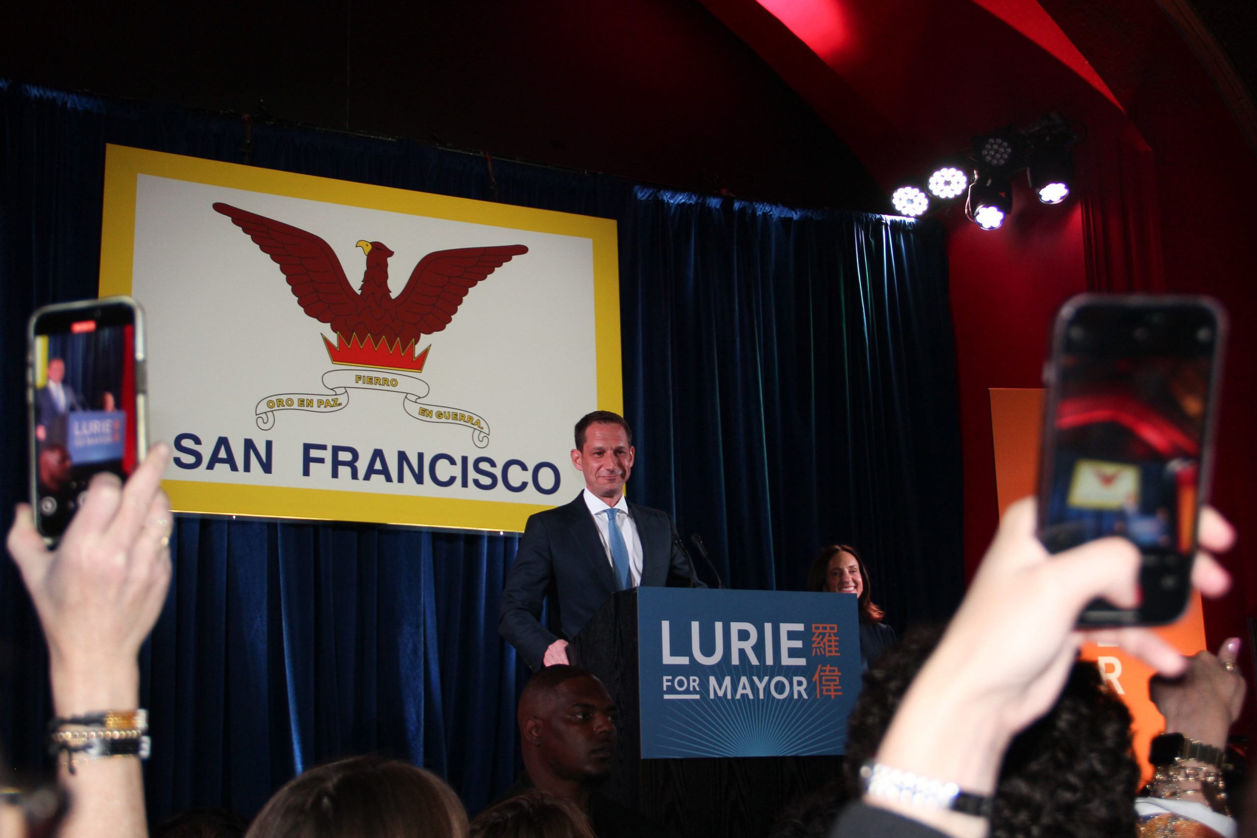 Person speaking at a podium labeled "Lurie for Mayor" with people holding phones. A large San Francisco emblem is displayed in the background.