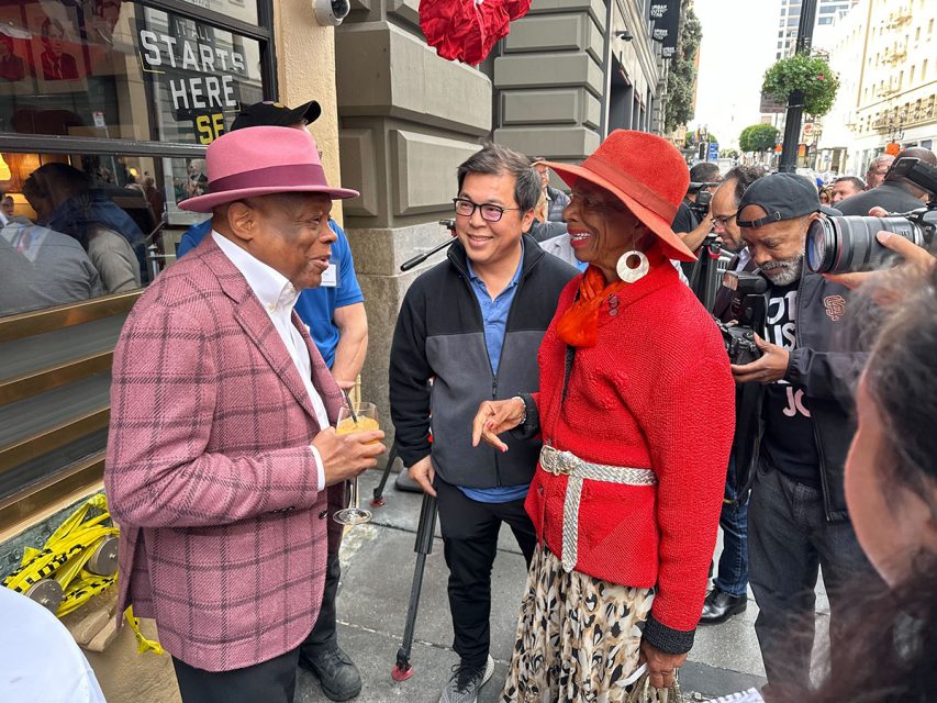 A group of people, including a man in a pink hat and a woman in a red outfit, engage in conversation on a bustling city sidewalk. Photographers are capturing the scene.