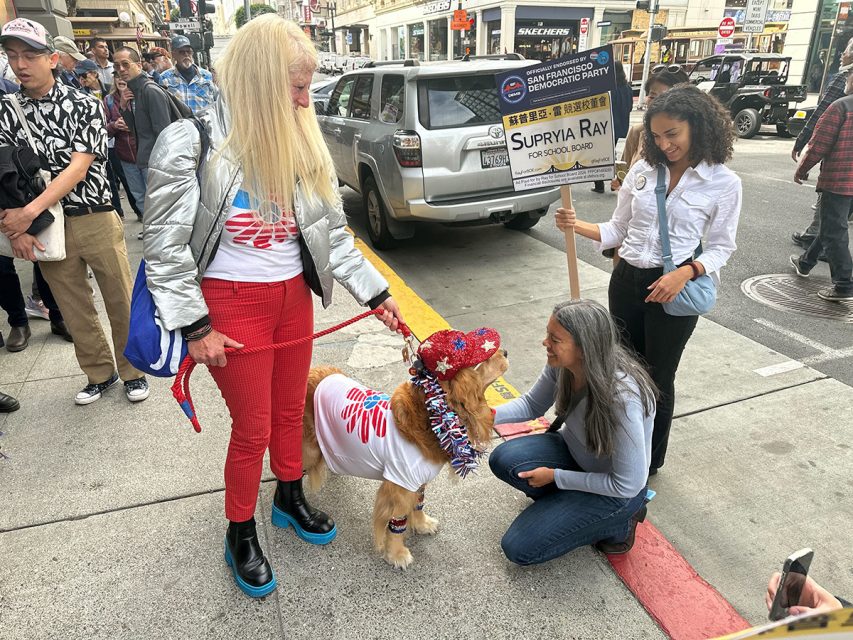 A group of people on a sidewalk with a dog in a decorative outfit. One person kneels to pet the dog, another holds a campaign sign.