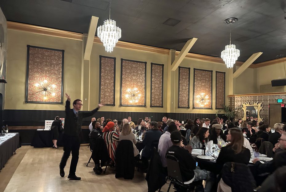 Mission Local managing editor Joe Eskenazi gestures energetically in front of a seated audience in an elegant room with chandeliers and patterned walls.