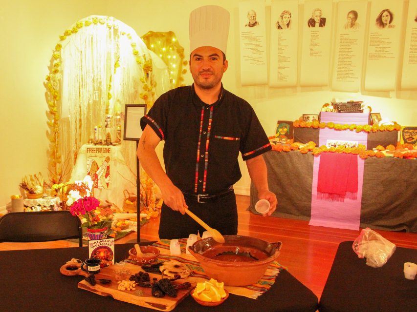 A chef in a tall hat prepares chocolate at a table with various ingredients. In the background, a decorated altar is visible, along with portraits on a wall.