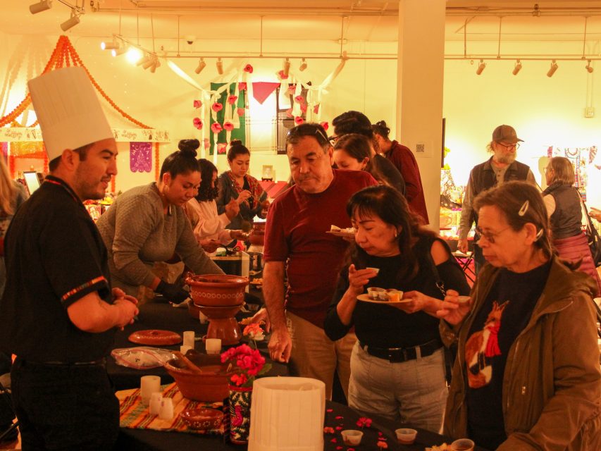 People gathered at a cultural food event, sampling dishes from a buffet-style setup. A chef in a tall hat serves food inside a warmly lit room.