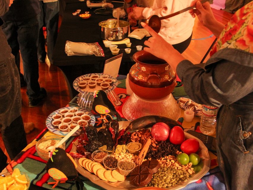 People preparing food at a buffet with a variety of ingredients and dishes, including a pot and a platter with vegetables, seeds, and crackers.