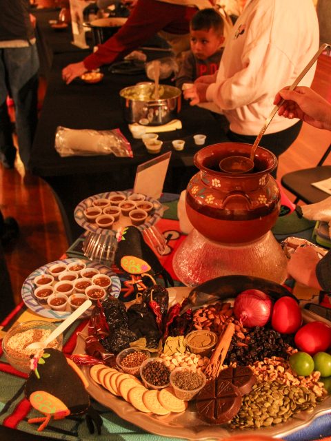 Table with assorted spices, chocolate, and ingredients, alongside a pot of melted chocolate. People are preparing food in the background. A child observes.