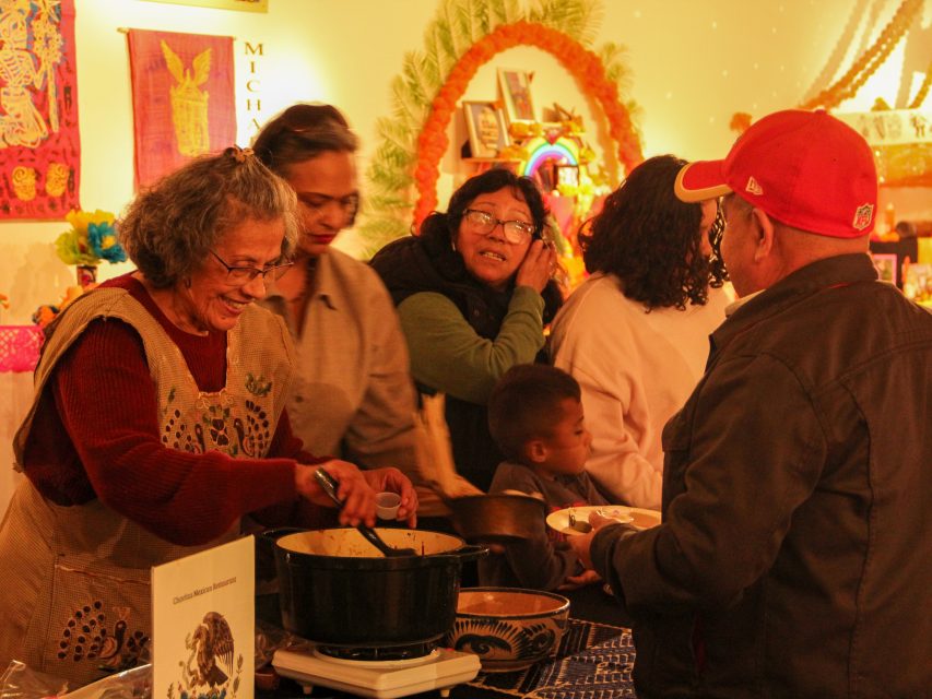 A woman serves food from a pot to people in line at a communal gathering. The setting is warm and decorated with colorful ornaments and art.