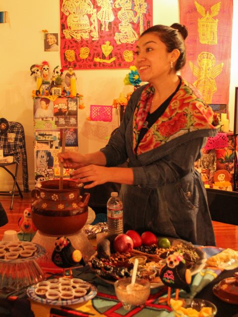 A woman stands at a table with traditional food and drinks. The background features colorful decorations and Day of the Dead motifs.