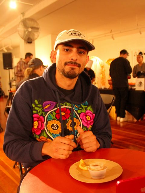 A man in a beige cap sits at a red table, holding utensils. He has a plate with sauce and a lemon slice. The room has wooden floors and several people in the background.