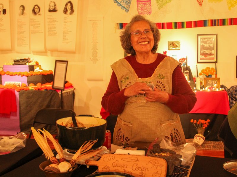 An elderly woman smiles while standing behind a table with traditional cooking ingredients and decorative items.