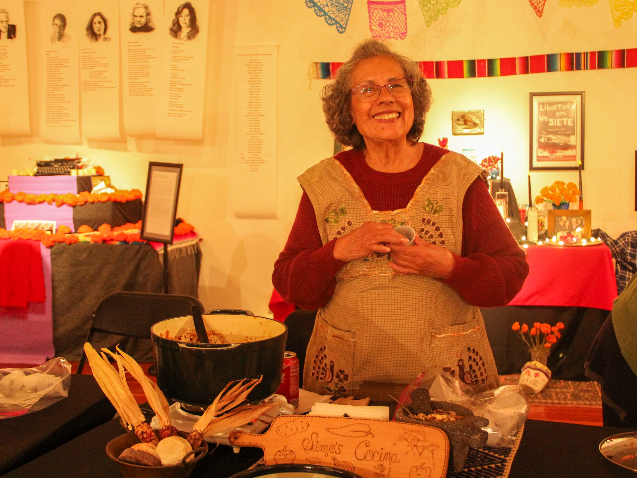 An elderly woman smiles while standing behind a table with traditional cooking ingredients and decorative items.