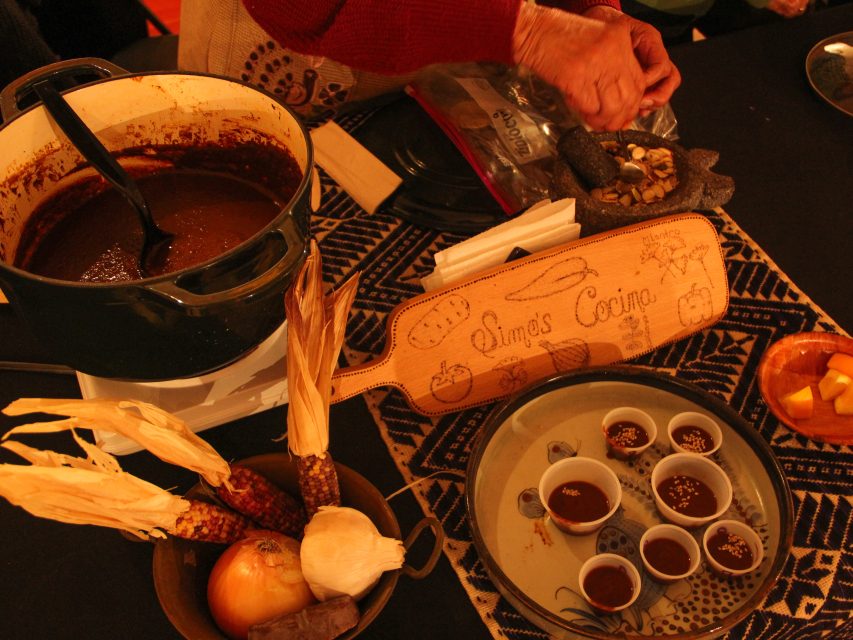 Cooking scene with a pot of sauce, spices, corn, and onion on a counter. A person prepares ingredients using a mortar. Various sauces are in small cups on a patterned tablecloth.