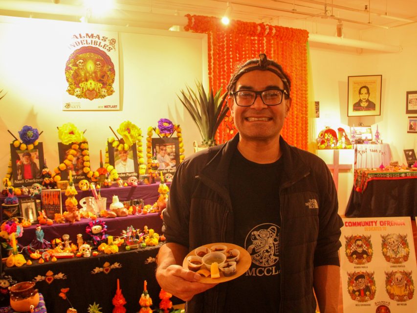 A person smiling and holding a plate of small cups in front of a Día de los Muertos altar decorated with flowers, candles, and colorful items.