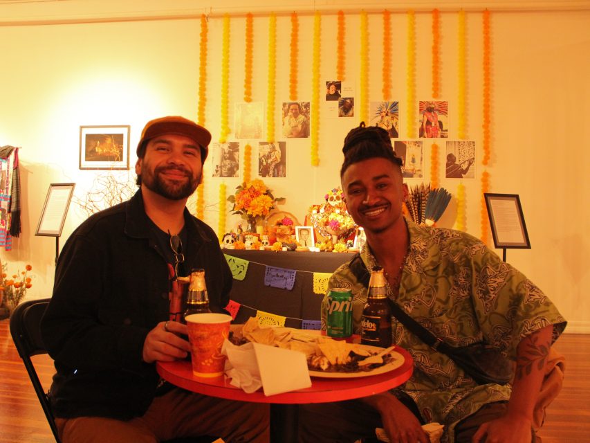 Two people sitting at a table with drinks and snacks, smiling at a festive indoor setting with decorations and photos on the wall behind them.