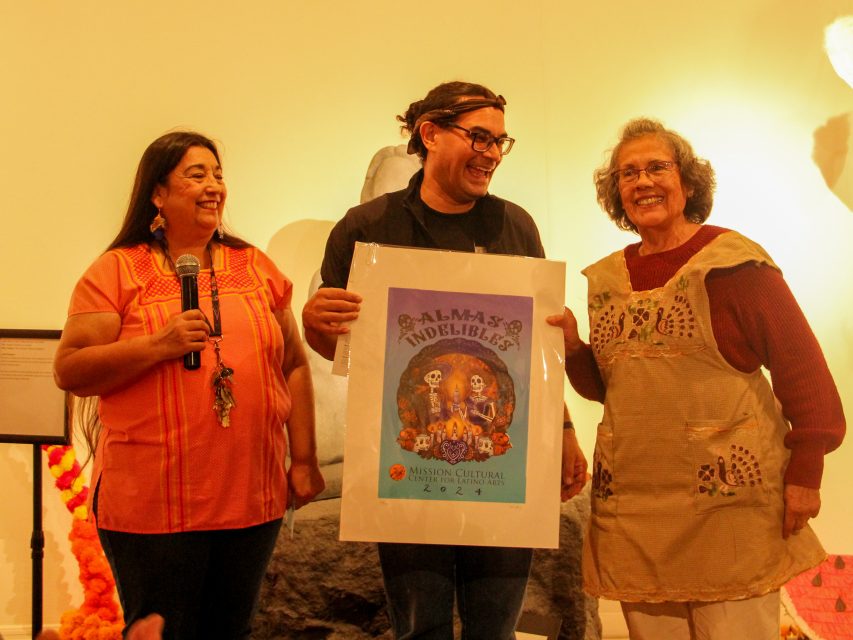 Three people are standing together, smiling. The middle person is holding a large poster with colorful artwork titled "Palmas Indebles." One person on the left is holding a microphone.