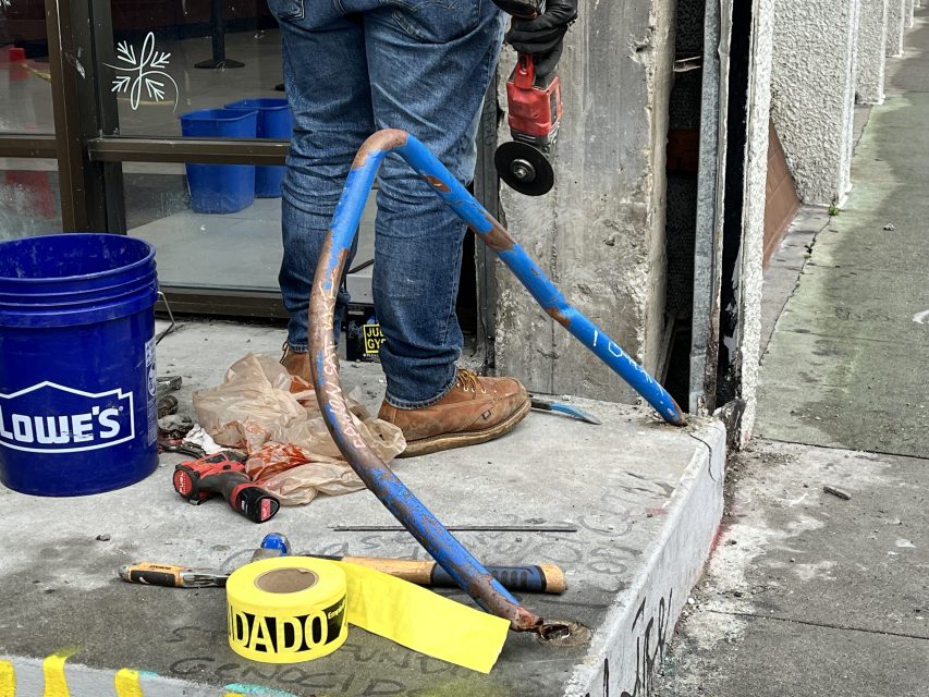 Person wearing jeans and boots working with a power tool on a concrete step. A bent blue pipe, tools, and tape are visible.