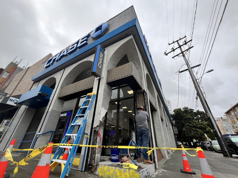 Construction work outside a Chase bank building with yellow caution tape, a blue ladder, traffic cones, and a worker in a gray hoodie.