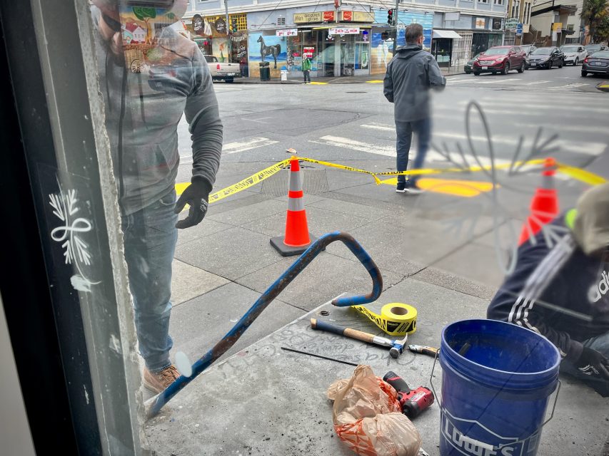 Workers repairing a sidewalk with tools and equipment, two traffic cones, and caution tape on a city street corner.