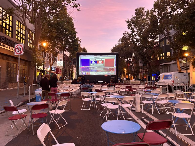 Outdoor movie screening set up on a street with rows of folding chairs facing a large screen displaying a game. The sky is pink, and trees line the street.