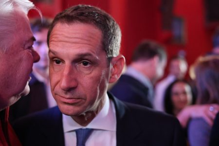 In a crowded room with red walls, a man in a suit and tie listens intently, his gaze unwavering as if absorbing the latest insights on the Trump discussions.