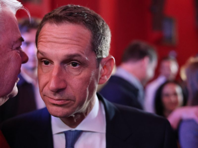 In a crowded room with red walls, a man in a suit and tie listens intently, his gaze unwavering as if absorbing the latest insights on the Trump discussions.