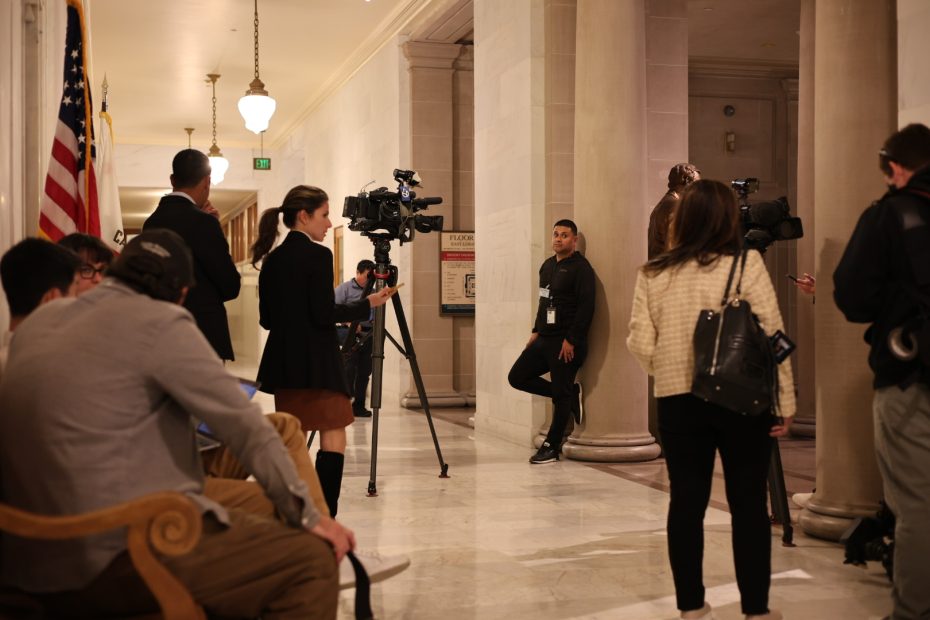 A group of journalists with cameras and microphones gather in a hallway with a U.S. flag and columns.