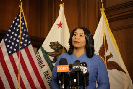 A woman stands at a podium with microphones in front of American, California, and city flags.