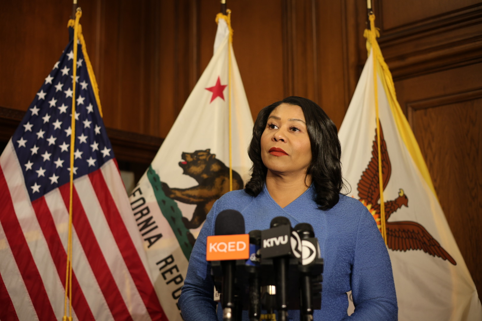 A woman stands at a podium with microphones in front of American, California, and city flags.