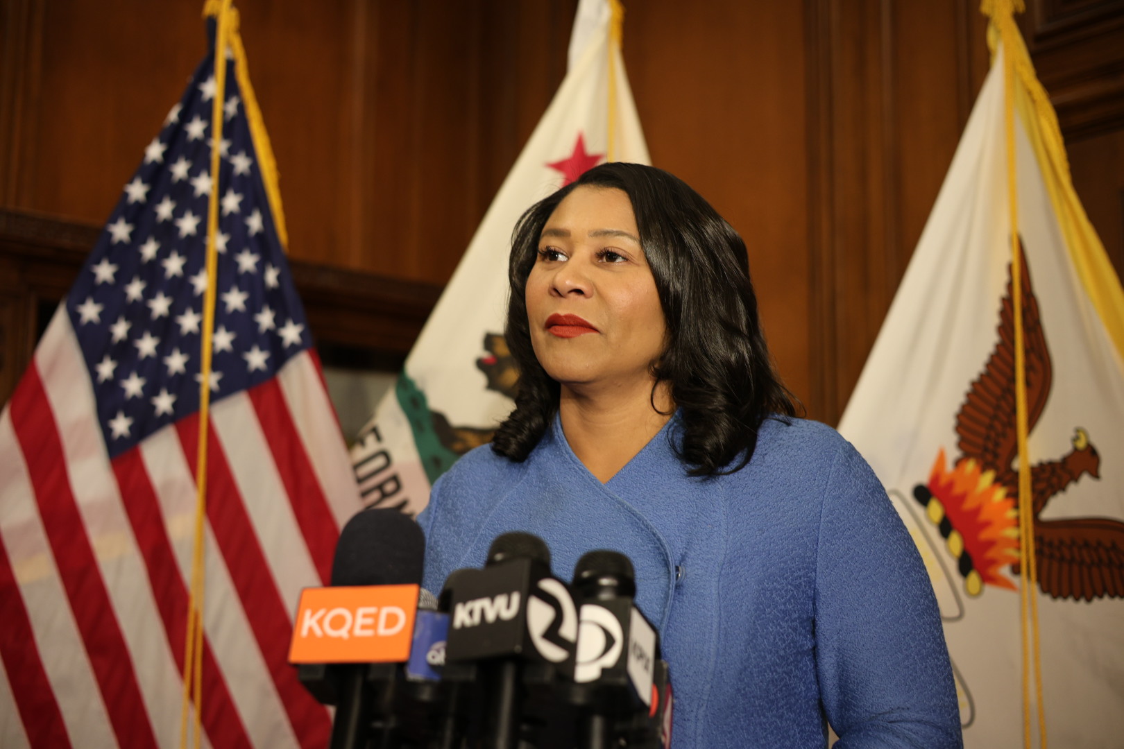 A woman speaking at a podium with microphones in front of U.S. and California flags.