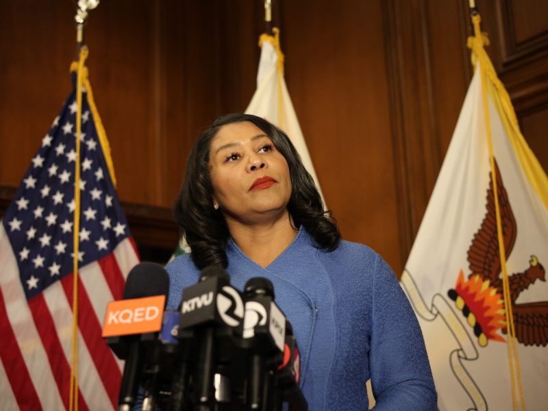 一位身著藍色服裝的女士站在講台前，面對多支麥克風，其中包括標有KQED和KTVU的麥克風，背景有多面旗幟。