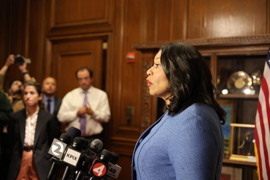 A woman in a blue suit speaks at a podium surrounded by microphones, with several people watching in the background inside a wood-paneled room.