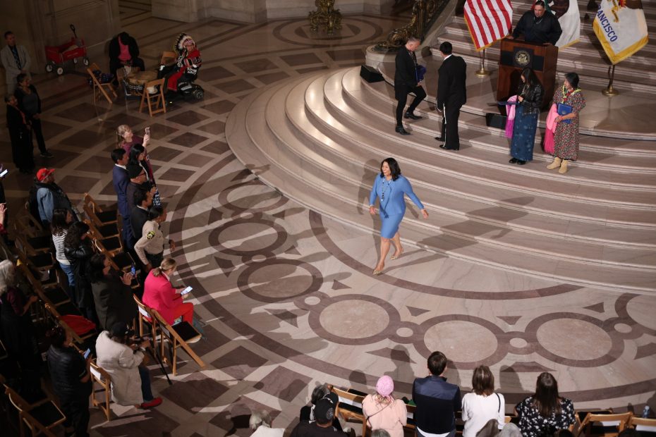 Person walking across a large, patterned floor towards a crowd and stage with officials and flags.