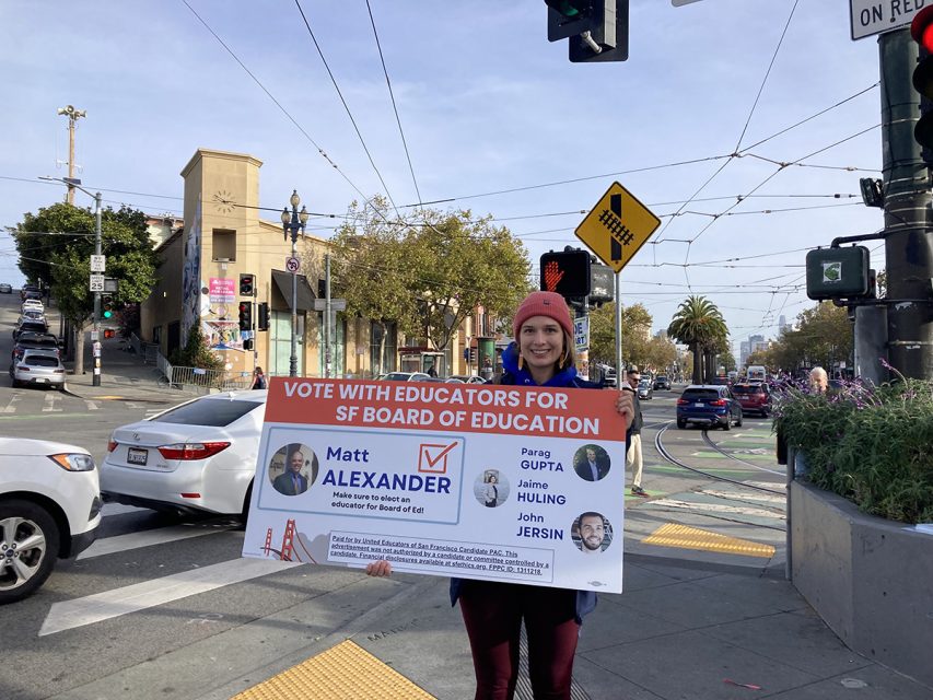 A person stands on a street corner holding a large sign advocating for a candidate in the SF Board of Education election.