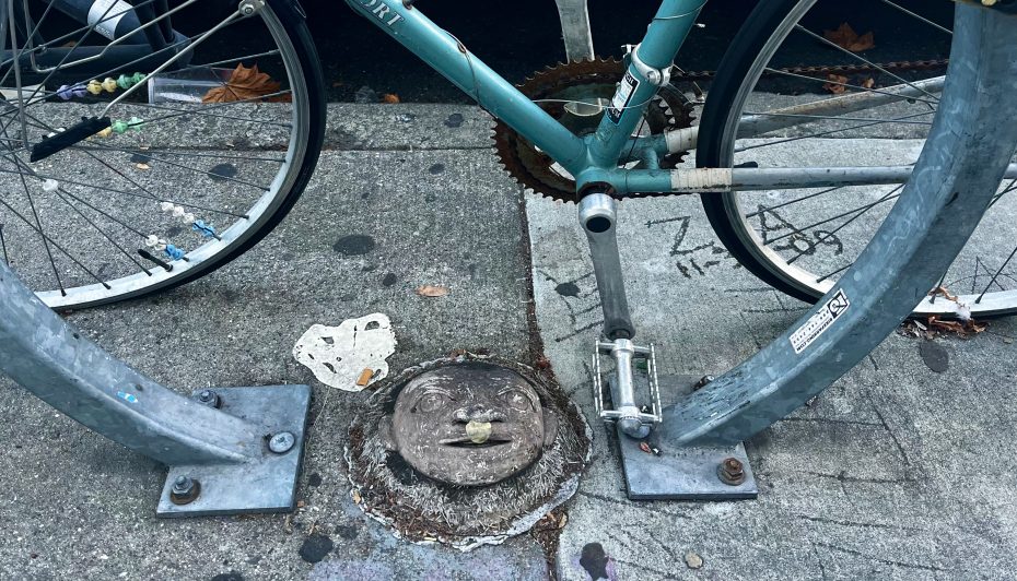Bicycle locked to a U-shaped metal bike rack on a sidewalk, with a face-like carving on a circular ground fixture.