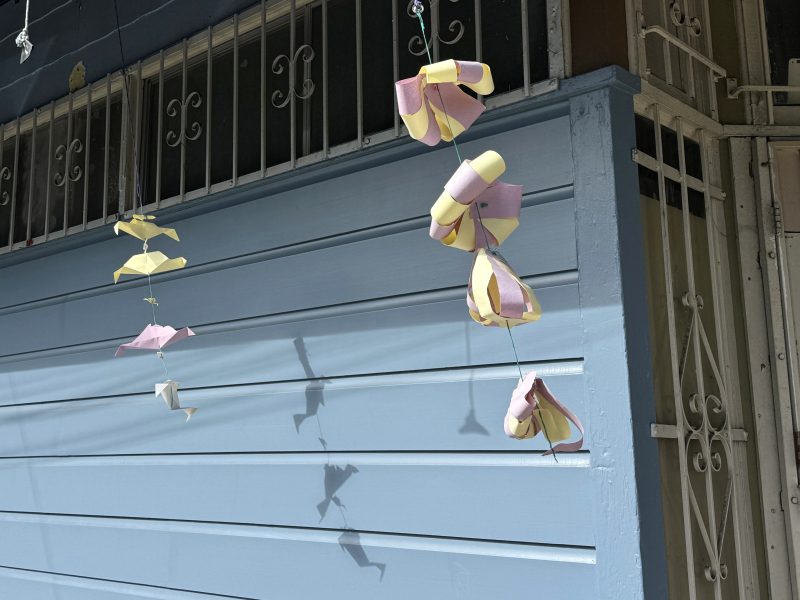 Colorful paper wind chimes hang in front of a pale blue wall with a metal grate window and door nearby. Shadows of the chimes are cast on the wall.