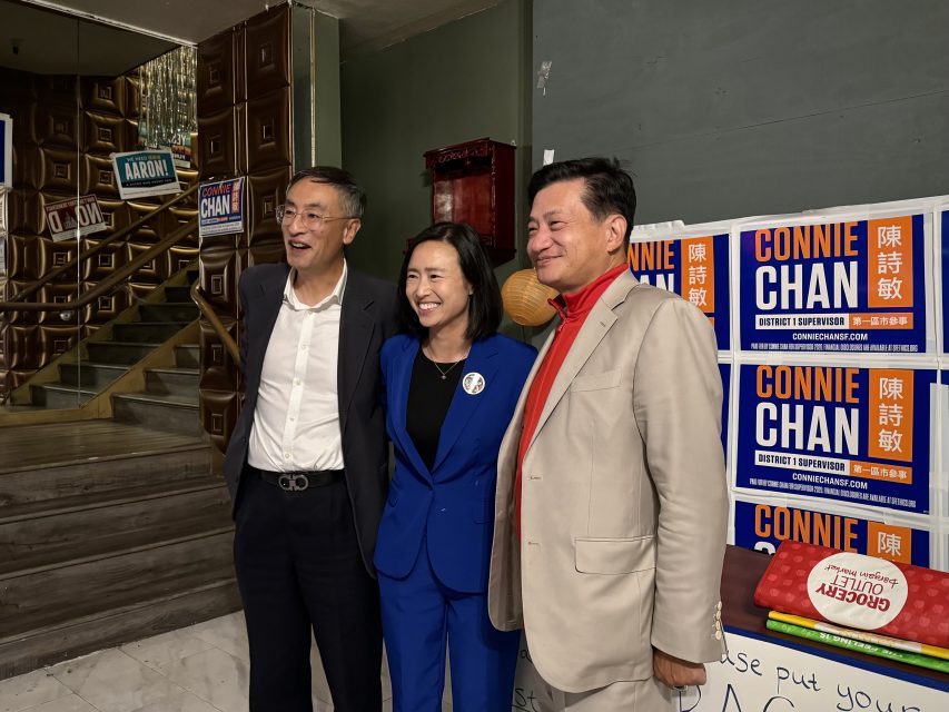 Three people stand together smiling in front of campaign signs in a room, with wooden stairs and a poster saying "Connie Chan.