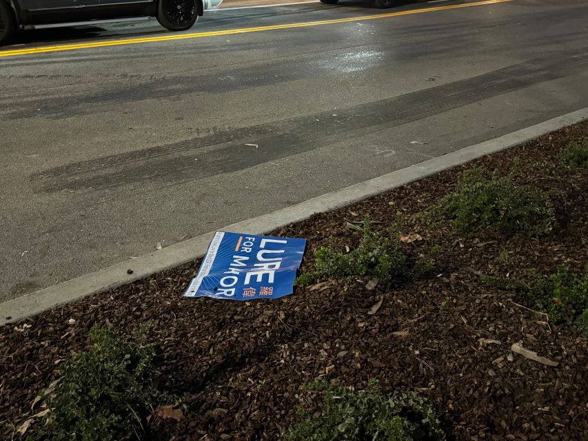 A "Lurie for Mayor" sign rests on the dirt beside a street with cars on it.