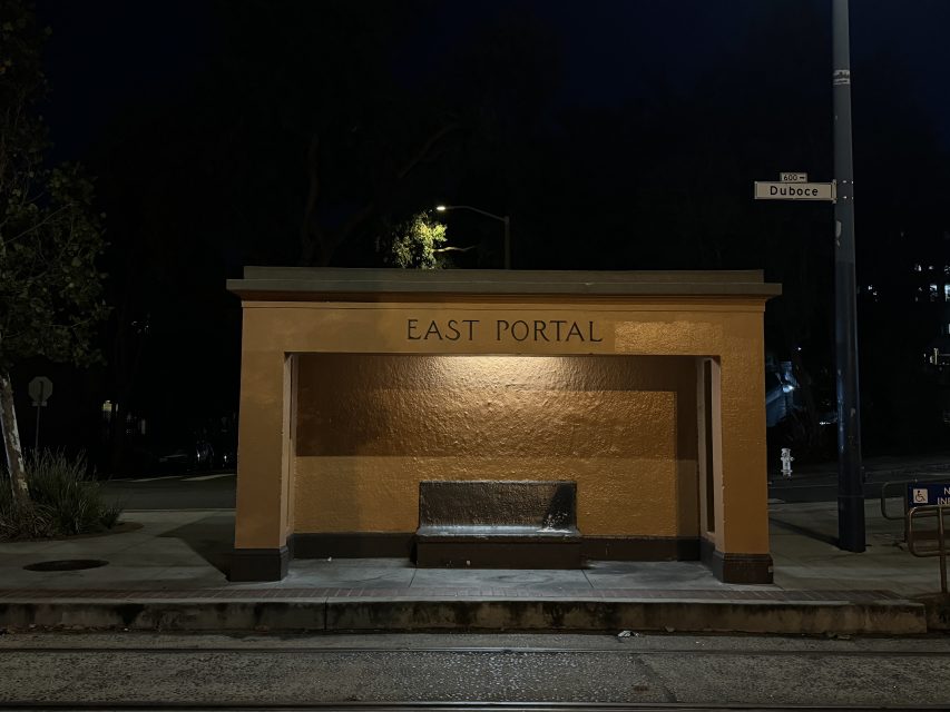 A small, orange building marked "East Portal" at night, with a dimly lit street and a street sign reading "Duboce."
