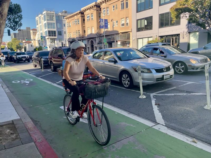 Person biking on a city street in a designated bike lane, wearing a helmet. Cars are visible in traffic lanes to the right. Buildings are in the background.