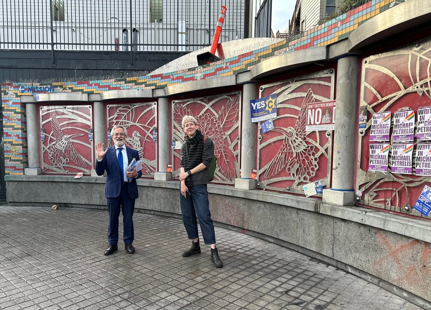 Two people stand in front of a mosaic wall adorned with red and white designs. One, in a suit, waves enthusiastically as if on the campaign trail for an election, while the other stands casually. Posters and signs peppered with text partially cover the vibrant backdrop.