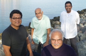 Four men are standing and sitting on rocks by a calm waterfront, with smiles and casual clothing.