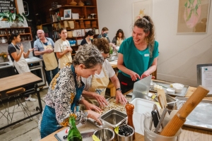 A group of people in a kitchen participates in a cooking class, with focus on two women preparing ingredients at a table, surrounded by culinary tools and a shelf with containers in the background.