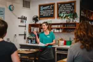 A woman in a teal shirt stands in a kitchen, holding a paper, speaking to a group. Behind her are chalkboards and shelves with kitchen items.
