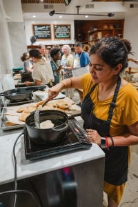 A woman in a yellow shirt and black apron fries food on a stovetop in a busy kitchen with people in the background.