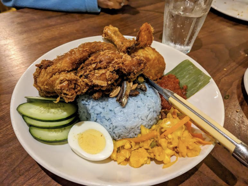 A plate of blue rice with fried chicken, cucumber slices, a boiled egg, vegetables, sambal, and a crispy garnish. A glass of water is in the background.