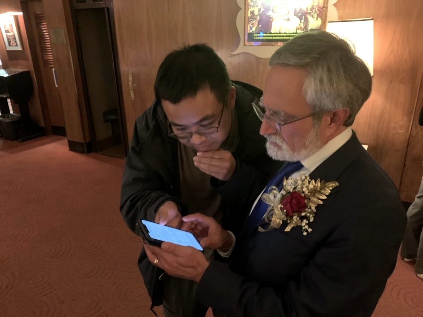 Two men look at a smartphone. The man on the right is holding the phone and wearing a suit with a boutonniere. They are standing in a dimly lit room.