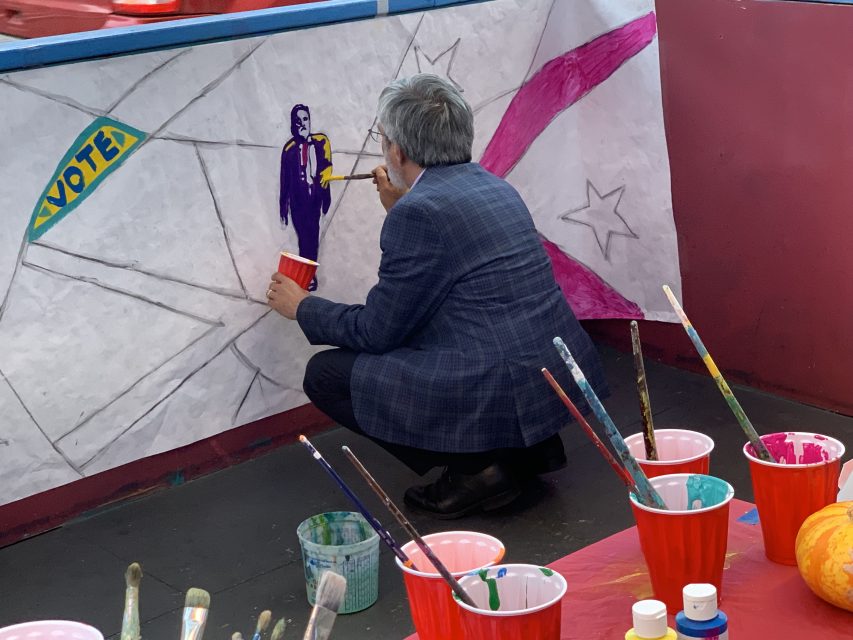 A person in a plaid jacket paints a mural with a figure and the word "VOTE" on a large sheet of paper. Several paintbrushes and cups with paint are in the foreground.