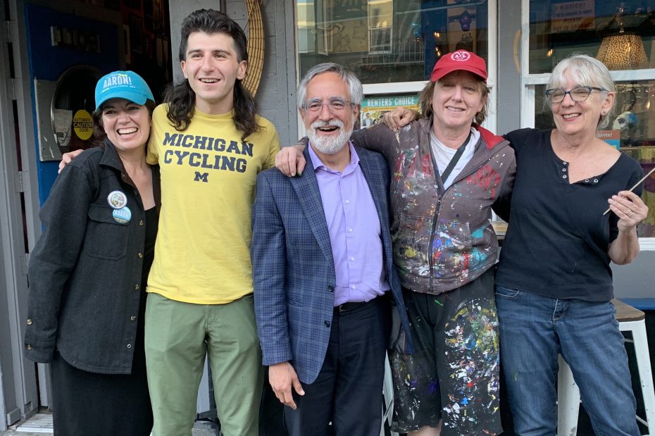 A group of five people stand closely together, smiling in front of a storefront. One person wears a "Michigan Cycling" shirt, and another is in a paint-splattered outfit.