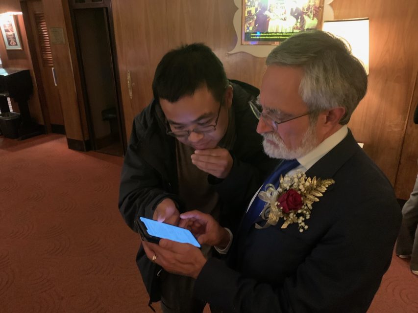 In a dimly lit room, two men focus intently on a smartphone screen. One, in glasses and a suit adorned with a boutonniere, discusses the election results while the other leans over to catch every detail illuminating from the device.
