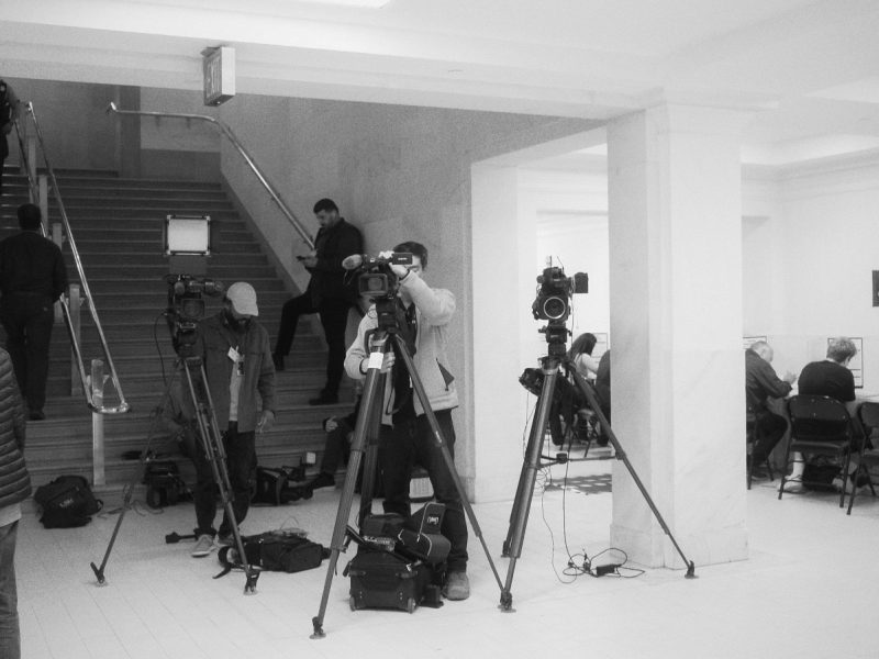 A group of people setting up video cameras in a hallway near a staircase. Others are seated at tables against the wall.