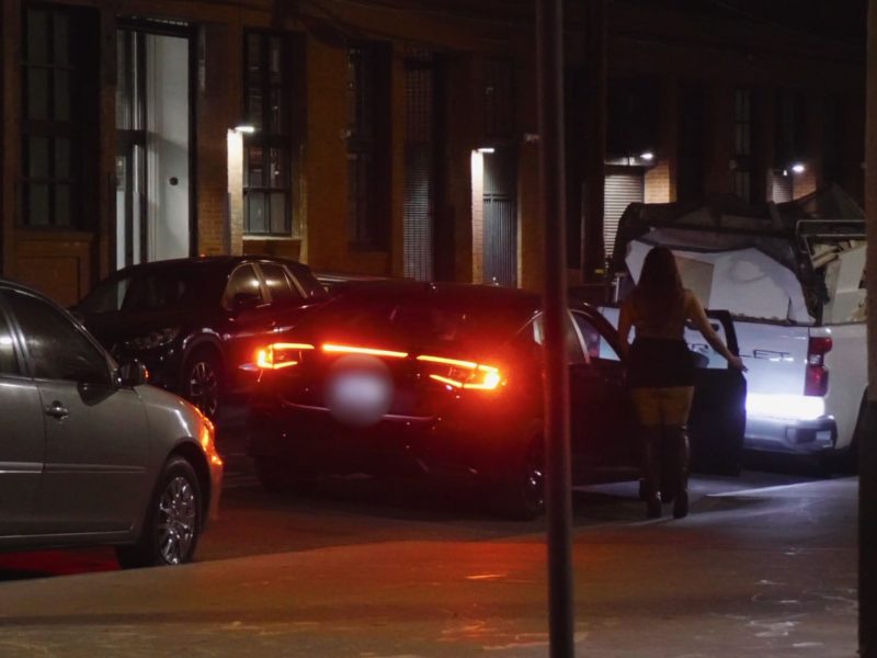 A woman walks on a dimly lit street towards a parked white vehicle. A black car with illuminated taillights is in the foreground.