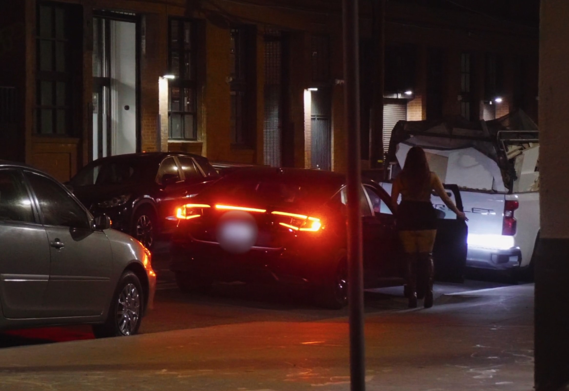 A woman walks on a dimly lit street towards a parked white vehicle. A black car with illuminated taillights is in the foreground.
